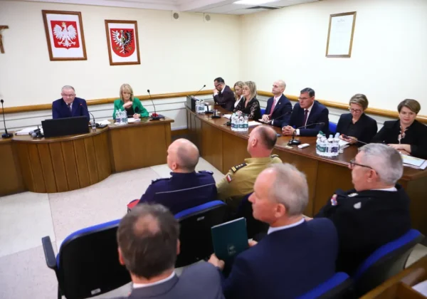 Uczestnicy V sesji Rady Powiatu w Płocku (kadencja 2024–2029) siedzą przy długim stole w sali konferencyjnej, z mikrofonami i wodą, w formalnych garniturach, a na ścianach wiszą herby i krzyż.