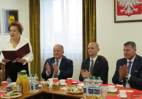 Kobieta w białej bluzce stoi i przemawia z czerwonym folderem w dłoni podczas XXVI Powiatowego Dnia Edukacji Narodowej, gdy siedzący w garniturach mężczyźni klaszczą przy stole z napojami i przekąskami.