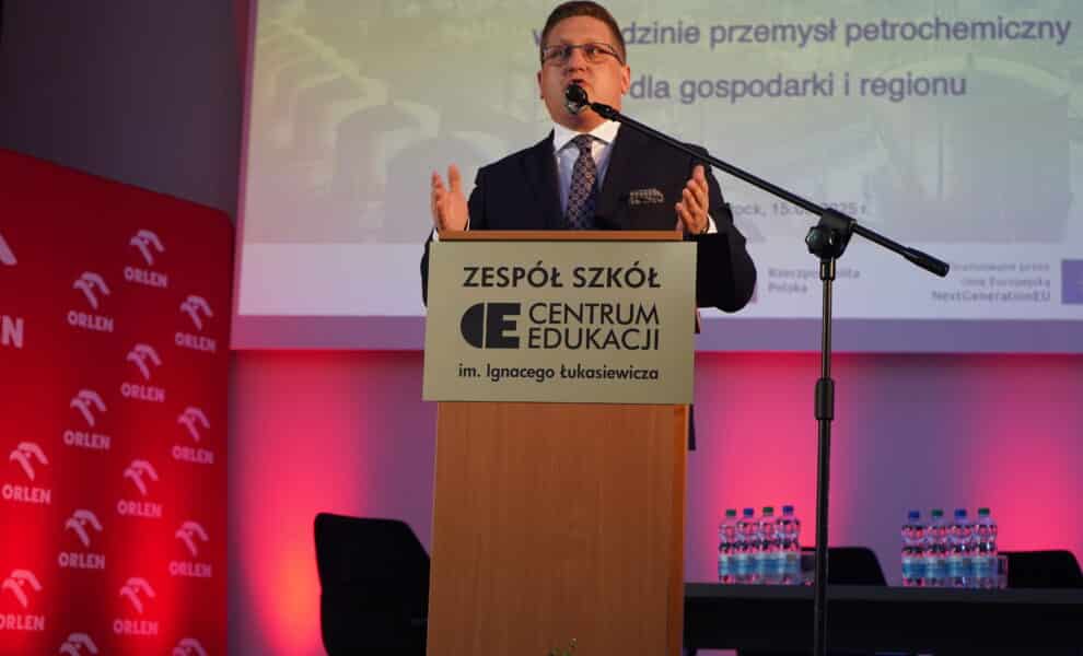 Mężczyzna w garniturze przemawia z mównicy na tle banerów związanych z branżą petrochemiczną.