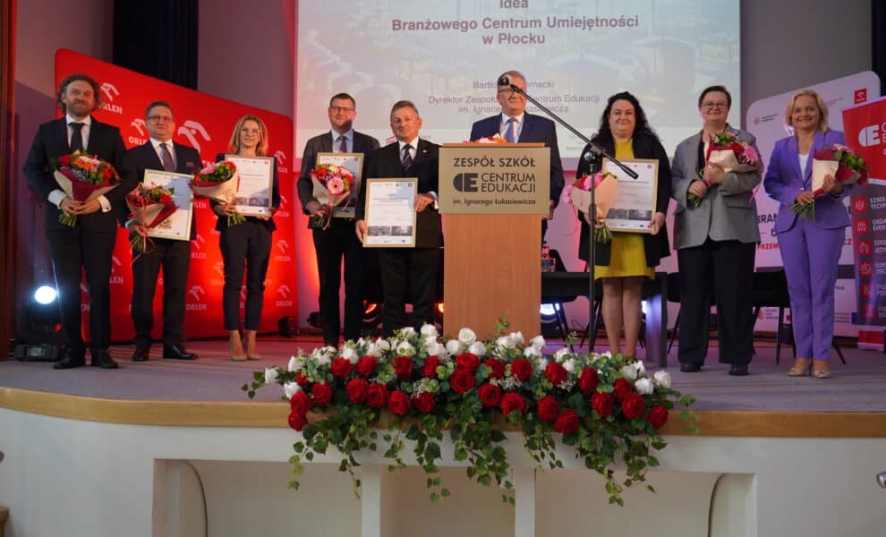 Na zdjęciu przedstawiono grupę osób stojących na scenie, które trzymają kwiaty i dyplomy, podczas wydarzenia promującego ideę Branżowego Centrum Umiejętności w Płocku.