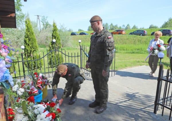 Dwaj żołnierze w mundurach wojskowych składają hołd przy pomniku, podczas obchodów rocznicy powodzi, a w tle widać uczestników ceremonii oraz alejkę z kwiatami.