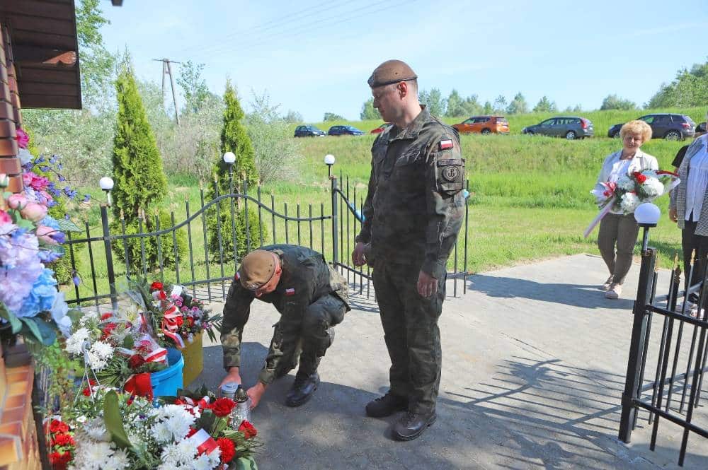 Dwaj żołnierze w mundurach wojskowych składają hołd przy pomniku, podczas obchodów rocznicy powodzi, a w tle widać uczestników ceremonii oraz alejkę z kwiatami.