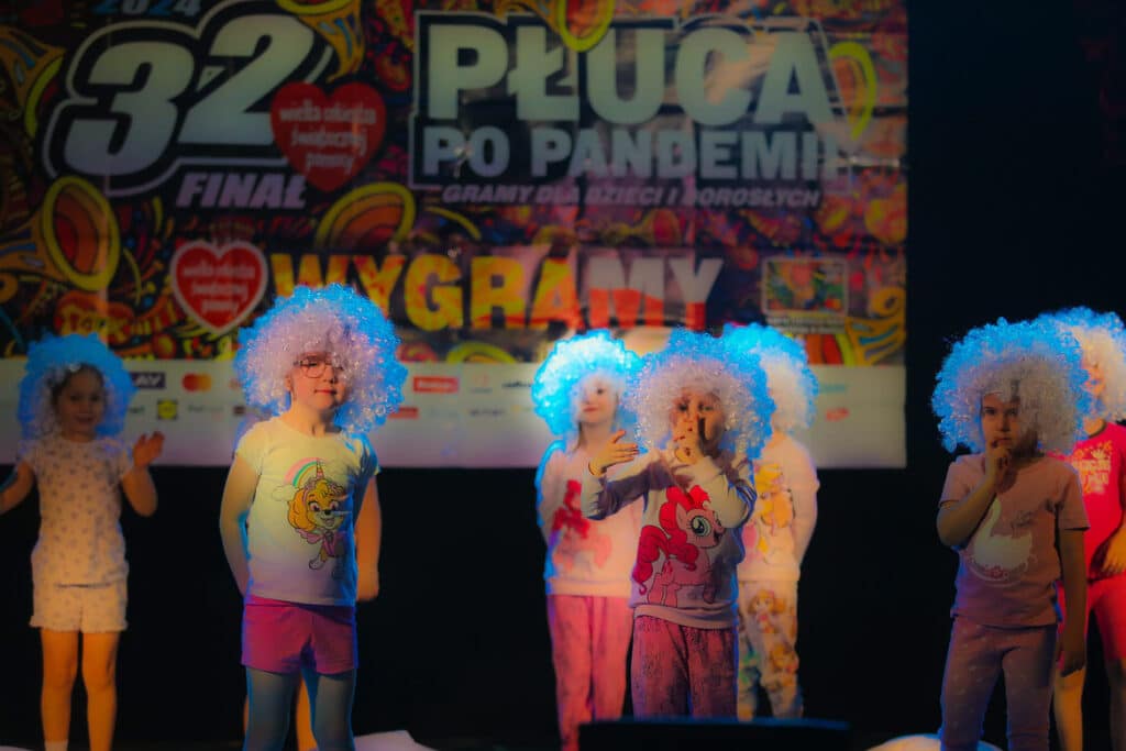 Na zdjęciu widoczna jest grupa dzieci w kolorowych, kręconych perukach, występująca na scenie podczas imprezy charytatywnej.