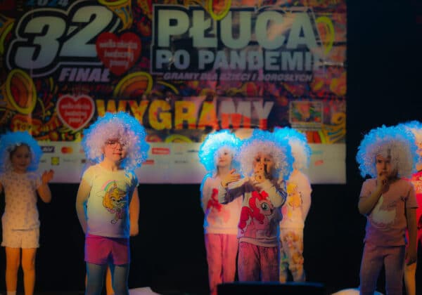 Na zdjęciu widoczna jest grupa dzieci w kolorowych, kręconych perukach, występująca na scenie podczas imprezy charytatywnej.