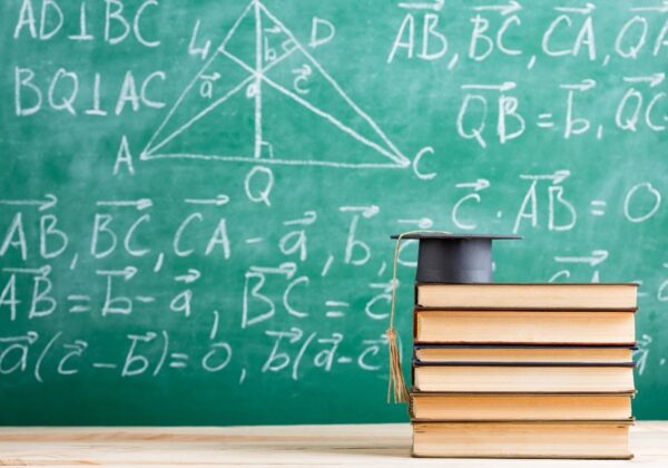 Na zdjęciu widać stos książek z czapką absolwenta na górze, przed zieloną tablicą pokrytą matematycznymi wzorami i rysunkami.