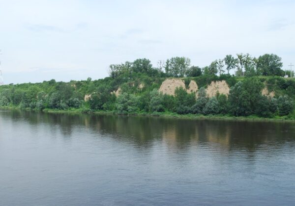 Widok na spokojną rzekę otoczoną zielonymi brzegami i urwiskami, z delikatnym odbiciem w wodzie.