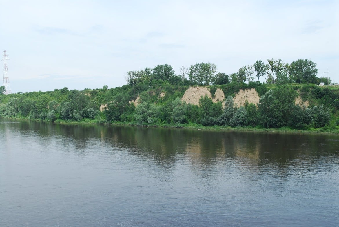 Widok na spokojną rzekę otoczoną zielonymi brzegami i urwiskami, z delikatnym odbiciem w wodzie.