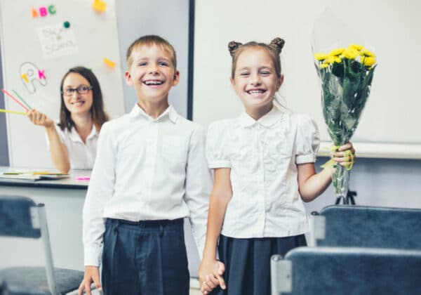 Dwoje uśmiechniętych dzieci w białych koszulach trzymających się za ręce, a dziewczynka w drugiej ręce trzyma bukiet żółtych kwiatów, podczas gdy nauczycielka w tle pokazuje kolorowe przybory szkolne.