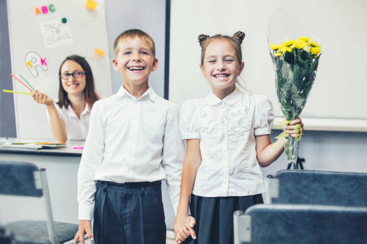 Dwoje uśmiechniętych dzieci w białych koszulach trzymających się za ręce, a dziewczynka w drugiej ręce trzyma bukiet żółtych kwiatów, podczas gdy nauczycielka w tle pokazuje kolorowe przybory szkolne.