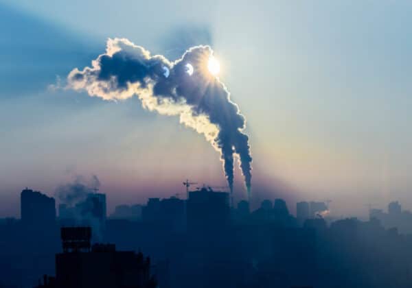 Zdjęcie przedstawia miejską panoramę z widocznymi kominami emituącymi dym w blasku wschodzącego słońca, tworzącym zjawiskową i tajemniczą atmosferę.