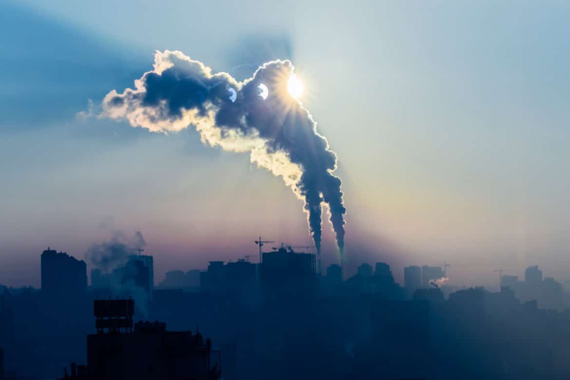 Zdjęcie przedstawia miejską panoramę z widocznymi kominami emituącymi dym w blasku wschodzącego słońca, tworzącym zjawiskową i tajemniczą atmosferę.