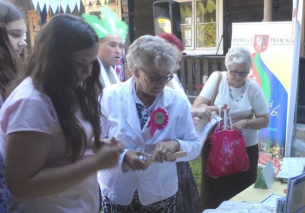 Na zdjęciu widać grupę ludzi uczestniczących w wydarzeniu promującym zdrowie, w którym kobieta w białym fartuchu analizuje materiały, a otaczające je osoby aktywnie uczestniczą w różnorodnych działaniach.