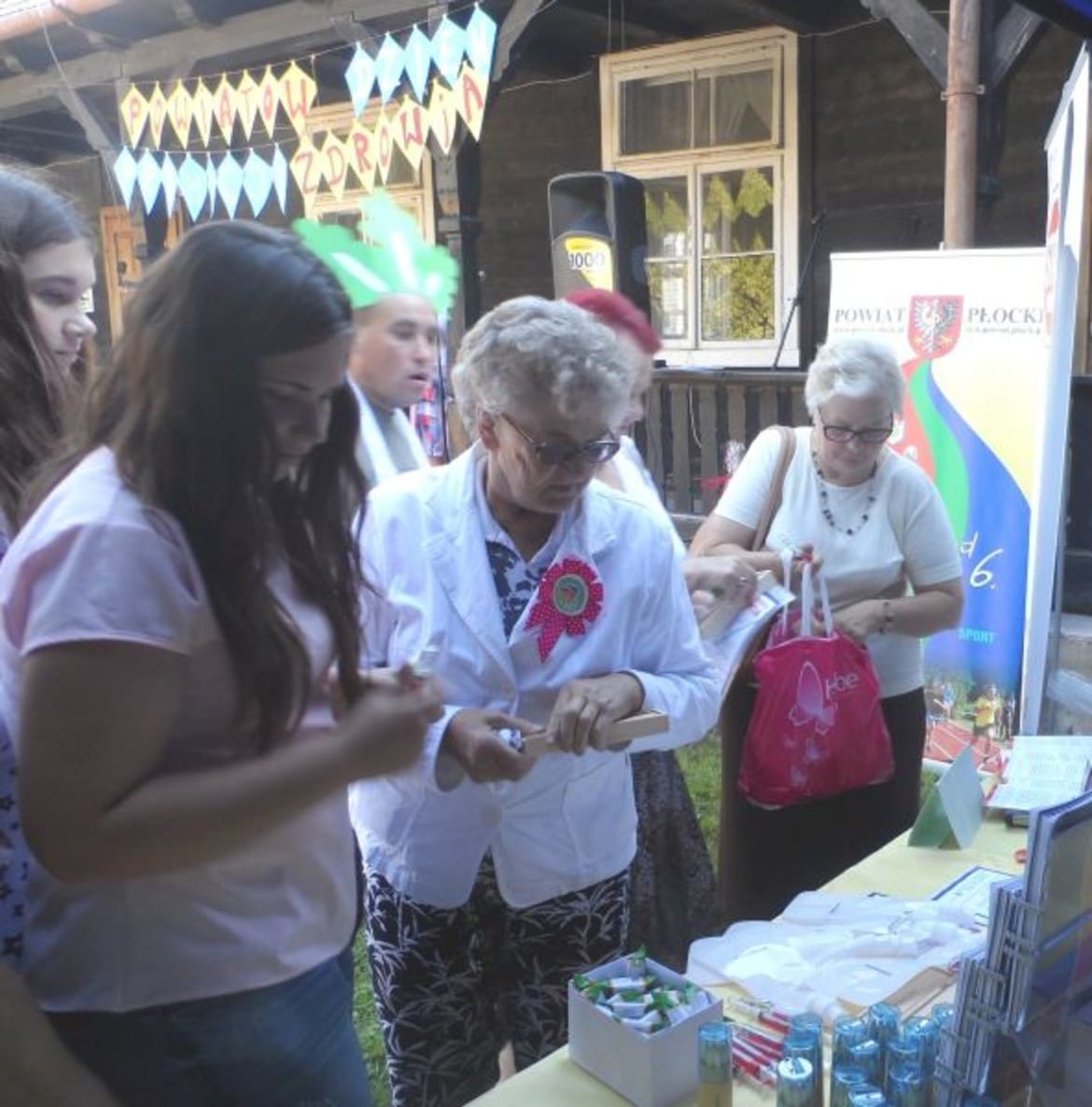 Na zdjęciu widać grupę ludzi uczestniczących w wydarzeniu promującym zdrowie, w którym kobieta w białym fartuchu analizuje materiały, a otaczające je osoby aktywnie uczestniczą w różnorodnych działaniach.