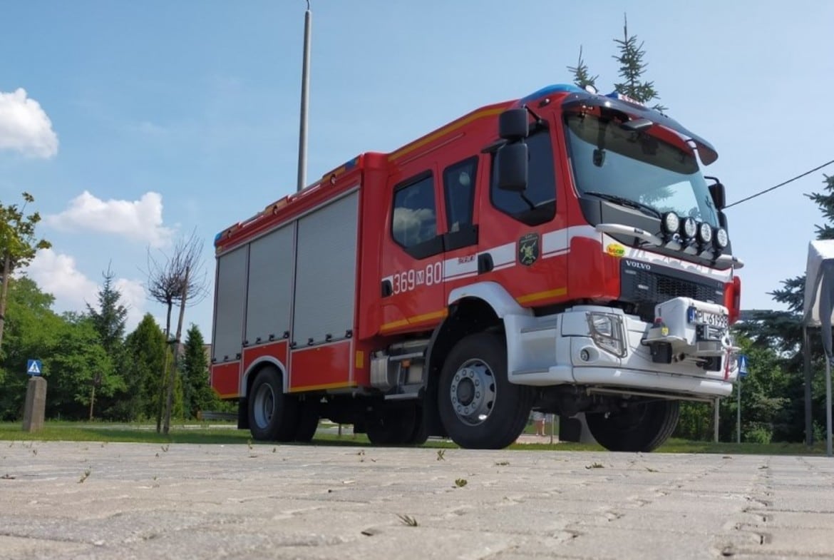 Na zdjęciu widoczny jest nowoczesny wóz strażacki w kolorze czerwonym zaparkowany na placu w otoczeniu zieleni i błękitnego nieba.