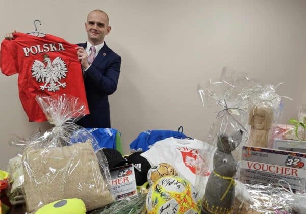 Na zdjęciu mężczyzna w garniturze trzyma czerwoną koszulkę z napisem "POLSKA" oraz otacza go stół z różnymi darami i gadżetami, w tym odzieżą i voucherami.