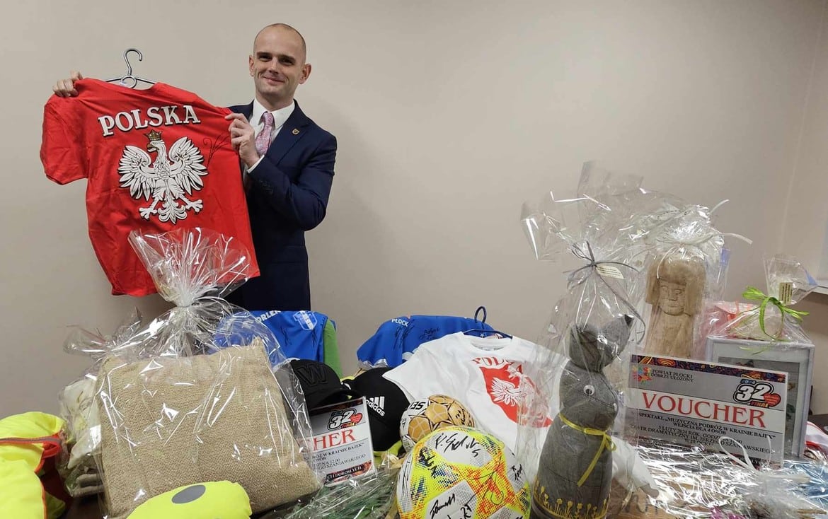 Na zdjęciu mężczyzna w garniturze trzyma czerwoną koszulkę z napisem "POLSKA" oraz otacza go stół z różnymi darami i gadżetami, w tym odzieżą i voucherami.