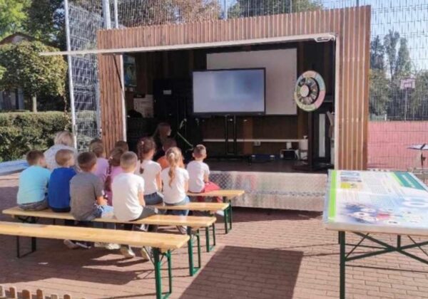 Na zdjęciu widać grupę dzieci siedzących na ławkach, które oglądają prezentację na ekranie w plenerowej scenerii.