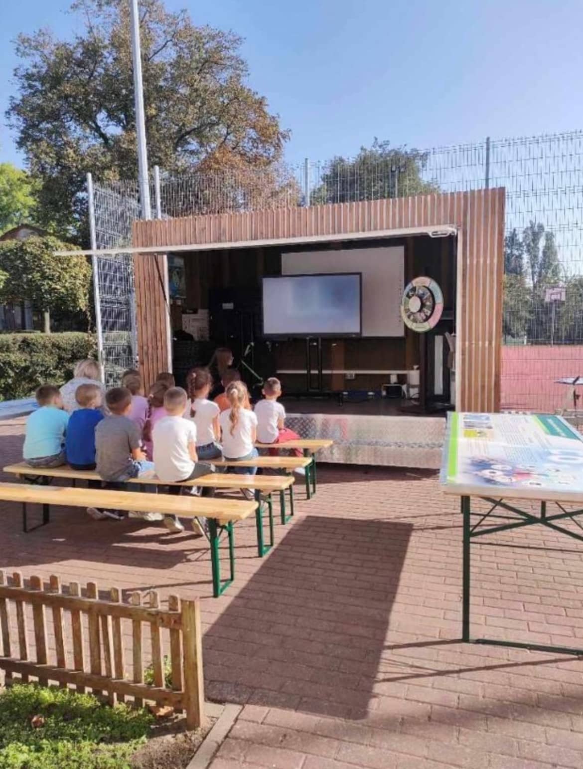 Na zdjęciu widać grupę dzieci siedzących na ławkach, które oglądają prezentację na ekranie w plenerowej scenerii.
