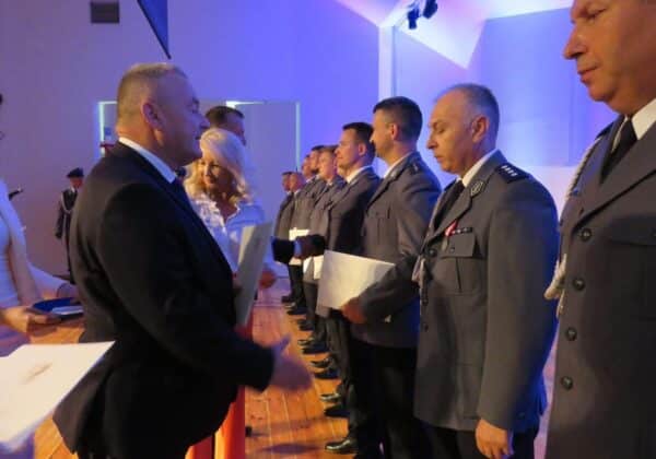 Na zdjęciu widoczna jest ceremonia wręczenia odznaczeń, w której uczestniczy grupa mężczyzn w mundurach oraz osoby w eleganckich strojach, prowadząca rozmowy i wręczająca dokumenty.