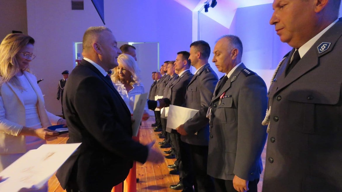 Na zdjęciu widoczna jest ceremonia wręczenia odznaczeń, w której uczestniczy grupa mężczyzn w mundurach oraz osoby w eleganckich strojach, prowadząca rozmowy i wręczająca dokumenty.