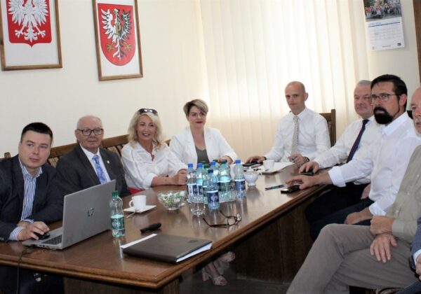 Grupa osób w formalnych strojach siedzi przy stole w sali konferencyjnej, na ścianie widoczne herbowe tarcze.