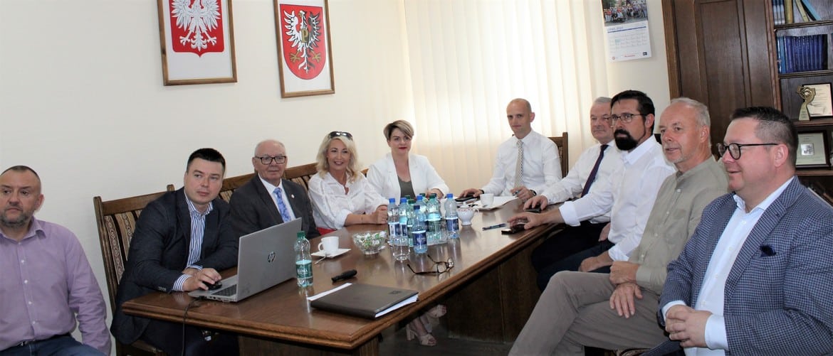 Grupa osób w formalnych strojach siedzi przy stole w sali konferencyjnej, na ścianie widoczne herbowe tarcze.