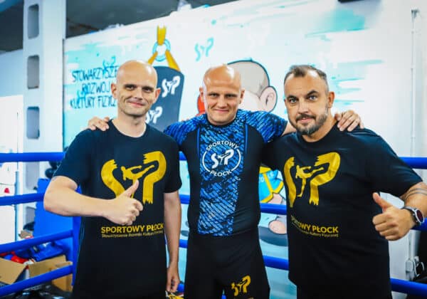 Trzech mężczyzn w koszulkach z logo "Sportowy Płock" pozują do zdjęcia w treningowej przestrzeni sportowej.