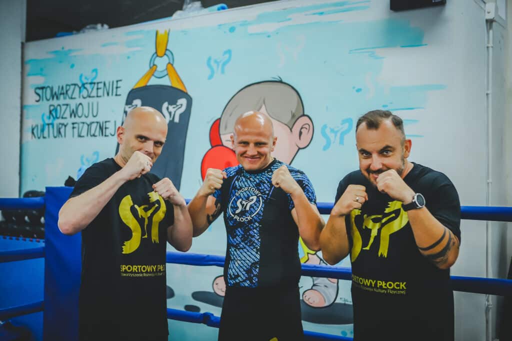 Trzej mężczyźni pozują do zdjęcia w sportowych koszulkach w sali treningowej, z muralem na tle przedstawiającym motywy sportowe.