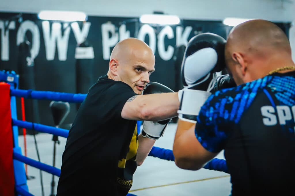 Zdjęcie przedstawia dwóch bokserów trenujących na ringu, skupionych na treningu w miejscu sportowym w Płocku.