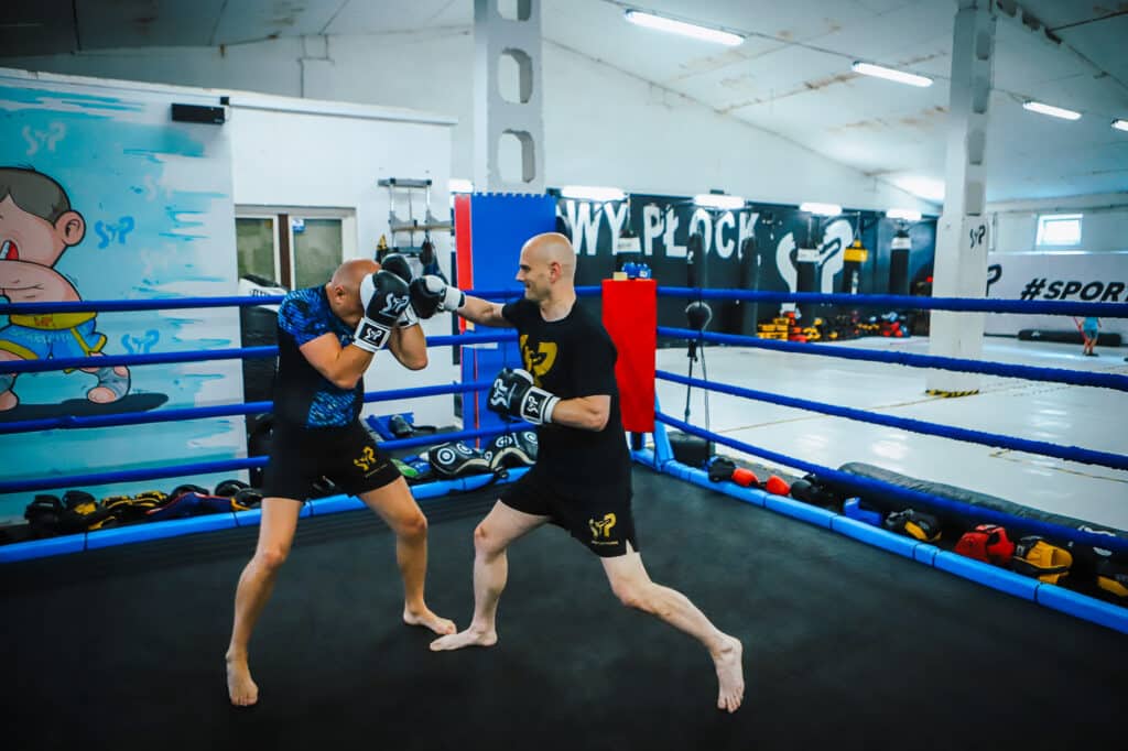 Na zdjęciu dwóch zawodników trenuje boks na ringu, pokazując zaangażowanie i pasję do sportu w nowoczesnej hali treningowej.