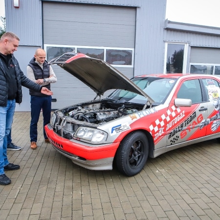 Dwaj mężczyźni oglądają silnik samochodu wyścigowego z otwartą maską na parkingu.