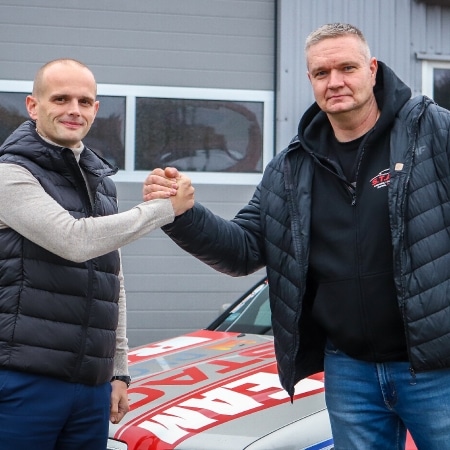 Na zdjęciu dwóch mężczyzn w sportowych ubraniach wita się, stojąc przed samochodem wyścigowym w garażu.