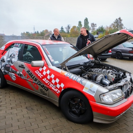 Na zdjęciu widoczny jest sportowy samochód z otwartą maską, przy którym rozmawiają dwaj mężczyźni, w tle inne pojazdy oraz jesienna sceneria.