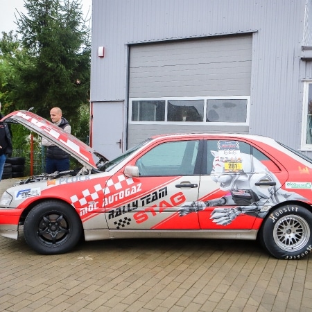 Na zdjęciu widoczny jest samochód wyścigowy z charakterystycznym malowaniem, stojący przed budynkiem, z otwartą maską oraz kilkoma osobami w tle.