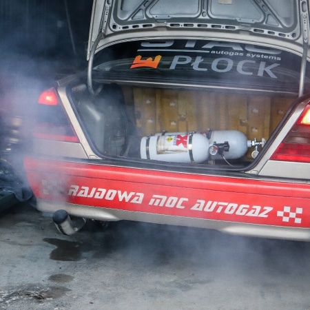 Z tyłu samochodu wyścigowego unosi się dym, a w bagażniku widoczny jest zbiornik autogazu z napisem „RAJDOWA MOC AUTOGAZ”.