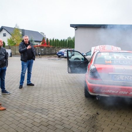 Na zdjęciu widać grupę mężczyzn obserwujących samochód, z którego unoszą się kłęby dymu, w tle widać parking i zabudowania.