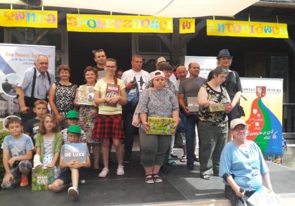 Zdjęcie przedstawia grupę osób stojących na scenie podczas Dnia Aktywnej Społeczności w Antoniówce, z transparentem w tle oraz różnorodnymi nagrodami.