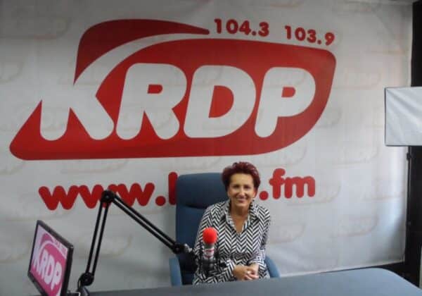 Na zdjęciu przedstawiona jest uśmiechnięta kobieta siedząca w studiu radiowym, z logo stacji KRDP w tle.