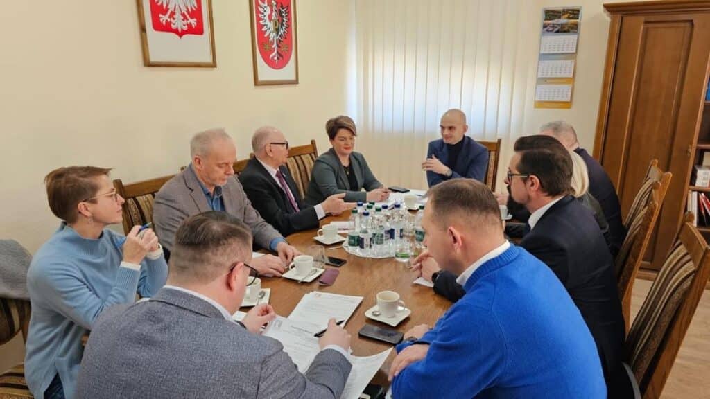 Na zdjęciu widoczna jest grupa osób siedzących przy stole w sali konferencyjnej, prowadzących dyskusję oraz analizujących dokumenty.