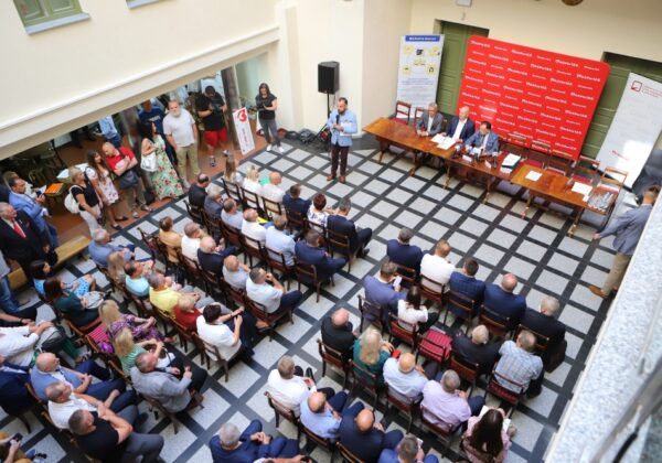 Zdjęcie przedstawia spotkanie w sali, na którym zgromadzeni uczestnicy słuchają wystąpienia prelegenta przy stole ustawionym na tle czerwonej tablicy z logo.