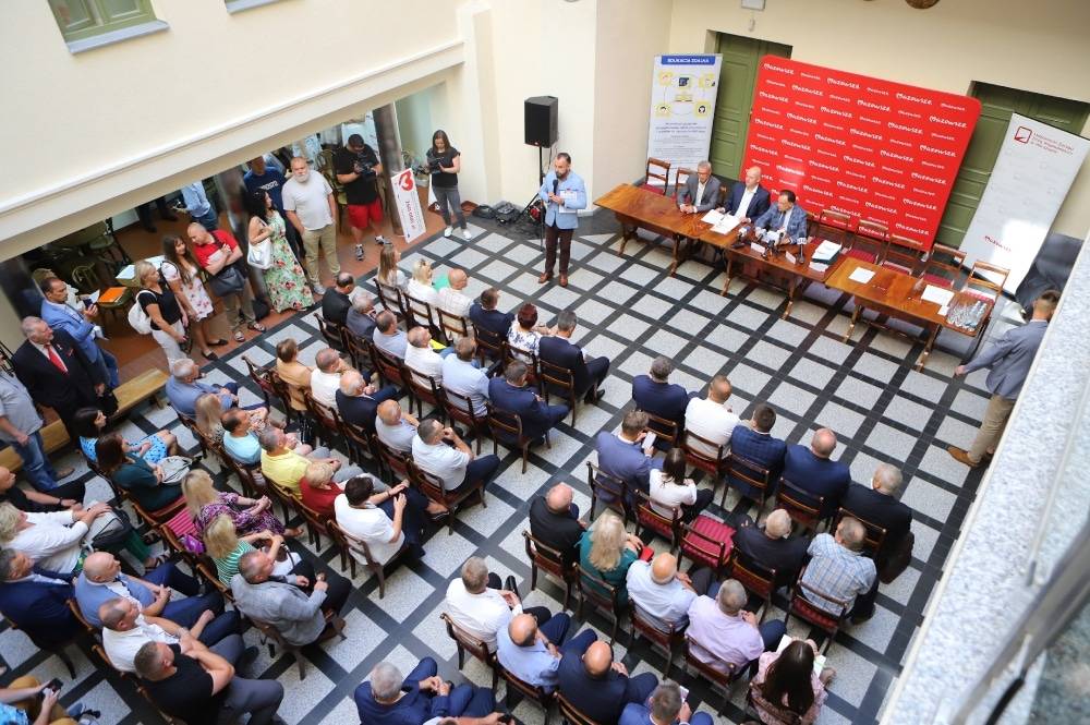 Zdjęcie przedstawia spotkanie w sali, na którym zgromadzeni uczestnicy słuchają wystąpienia prelegenta przy stole ustawionym na tle czerwonej tablicy z logo.