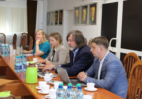 Na zdjęciu przedstawiono grupę osób uczestniczących w spotkaniu przy długim stole, z laptopami i napojami, w sali z portretami na ścianie.