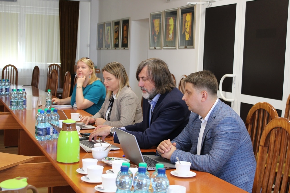 Na zdjęciu przedstawiono grupę osób uczestniczących w spotkaniu przy długim stole, z laptopami i napojami, w sali z portretami na ścianie.