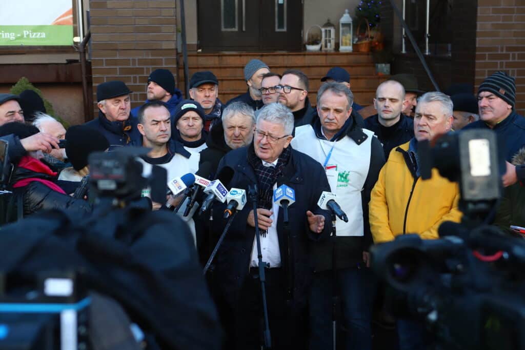 Na zdjęciu widoczna jest grupa mężczyzn, z których jeden przemawia do mikrofonów podczas konferencji prasowej, a pozostali słuchają go z zainteresowaniem.