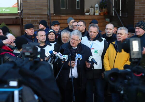 Na zdjęciu widoczna jest grupa mężczyzn, z których jeden przemawia do mikrofonów podczas konferencji prasowej, a pozostali słuchają go z zainteresowaniem.