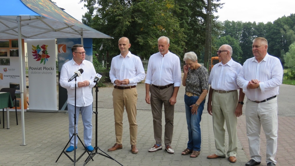 Na zdjęciu widoczna jest grupa osób stojących przed stoiskiem informacyjnym, przygotowujących się do ogłoszenia dotyczącego remontu drogi w Orszymowie.