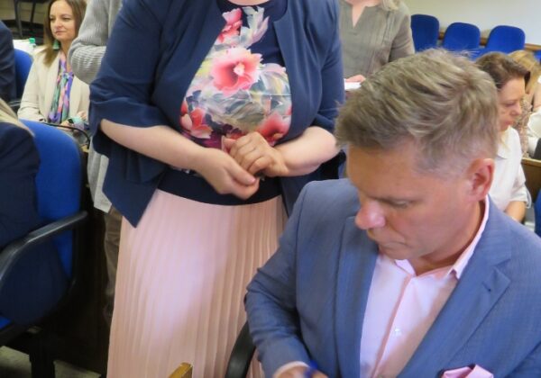 Na zdjęciu widoczna jest scena z wydarzenia, w którym uczestnicy czekają na podpis autora książki, siedzącego przy stole, podczas gdy jedna z osób z zainteresowaniem obserwuje proces podpisywania.