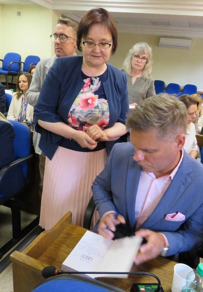 Na zdjęciu widoczna jest scena z wydarzenia, w którym uczestnicy czekają na podpis autora książki, siedzącego przy stole, podczas gdy jedna z osób z zainteresowaniem obserwuje proces podpisywania.