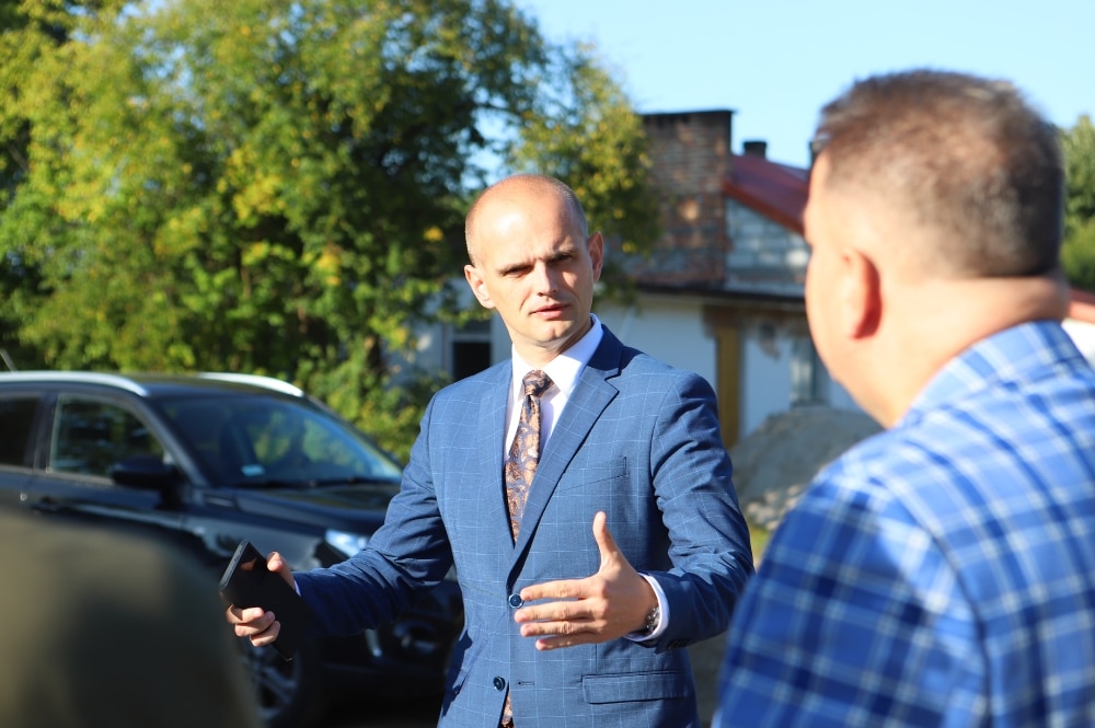 Na zdjęciu widać mężczyznę w garniturze, który gestykuluje i rozmawia z inną osobą, podczas gdy w tle znajdują się drzewa i samochód.