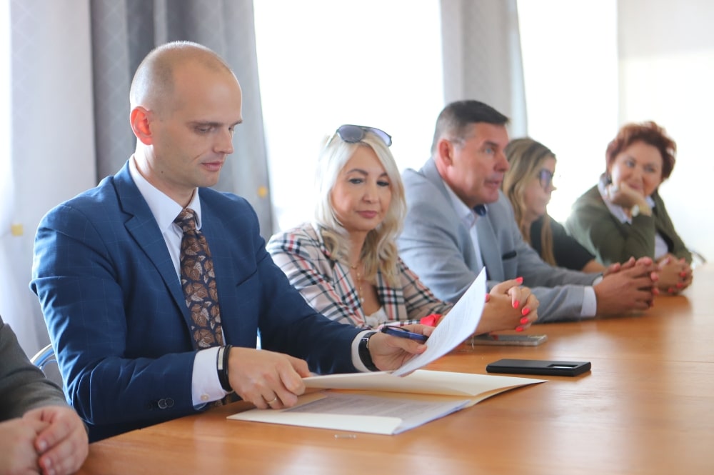 Na zdjęciu widać grupę osób siedzących przy stole, w trakcie spotkania, podczas którego jedna z osób przegląda dokumenty.